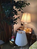 Photo shows two matching table lamps with pleated fabric lampshades placed on a wooden side table, surrounded by a faux ficus tree in a woven basket, two clocks, a yellow glass candy dish, and a plaster statue on the floor nearby.