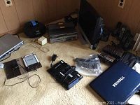 Wide shot showing Toshiba laptop, Sanyo DVD player, various cordless phones, Nook e-reader, and accompanying accessories on carpeted floor