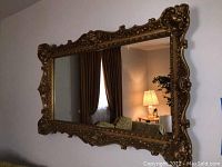 Full view of the large ornate wall mirror hung on cream colored wall showing decorative gold frame and reflection of the room.
