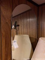 Mirror with a curved top, leaning against a wood-paneled wall, showing the front reflective surface and partial reflection of a lamp.