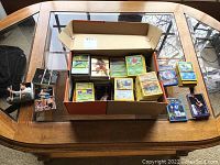 Large open cardboard box on a table containing multiple rubber-banded stacks of assorted Pokémon cards, a few small stacks of basketball and football cards beside the box