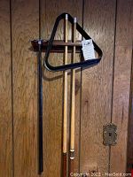Wide view of three wooden pool cues, black triangle rack and wall-mounted wooden cue rack with green felt rests, against wood panel wall.