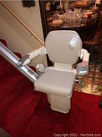 Front view of beige stair lift chair mounted on straight silver aluminum rail on red-carpeted staircase