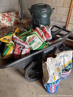 Wide view of Rubbermaid utility cart with multiple bags of garden soil, lawn care products, and two stacked green Rubbermaid trash cans with lids in the background.