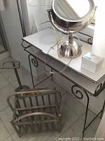 Bathroom vanity table with white top and black metal frame, metal wire wastebasket, chrome countertop mirror, soap bar, and paper towel roll on top, with a rustic metal magazine rack and metal toilet paper stand on floor beside it.