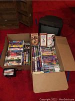 Two cardboard boxes filled with assorted VHS tapes placed on red carpet floor next to a black Quasar TV/VCR combo model VV-1309 unit.