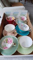 Six teacup and saucer pairs arranged in a box showing various colors and floral designs