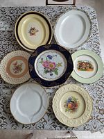 Seven assorted china dinner plates with a variety of floral, scenic, and embossed fruit patterns