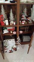 Bookshelf display with multiple Christmas decorations including resin trees, glass tree, ceramic figurines, glass vase, red clock and box of ornaments