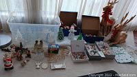 Wide photo showing all the assorted Christmas decorations including glass trees, reindeer statues, figurines, and boxes of ornaments on a table.