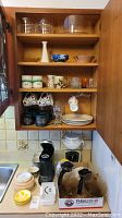 Photo of kitchen cabinet and countertop showing mugs, glasses, plates, K-Cup brewer, coffee carafes, and household scale.