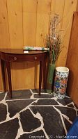 Half moon wooden hallway table with single drawer, tall green vase containing dried decorative grass, and tall ceramic umbrella stand with floral design.