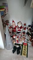 A wide view showing a collection of vintage Christmas decorations including several Santa figures, nutcrackers, small figurines, a silver cone tree, gift wrap rolls, and a green bow.