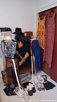 Photo showing vintage floral dresses hanging and multiple handbags on chairs and floor, with hand-stitched dragonfly scarves draped over chairs along with slippers on floor.