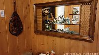 Full view of the vintage solid wood mirrored shelf mounted on wood-paneled wall, showing contents inside and carved paddle hanging next to it.