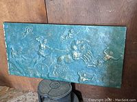 Full view of the large rectangular plaster bas relief wall plaque displayed against a wooden wall and resting above a stove. The relief depicts a dramatic neoclassical scene with horses, a chariot, and human and angelic figures in raised detail.