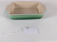 Angled front view of rectangular green Le Creuset baking dish with cream interior and side handles.