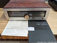 Front angled view of Luxman T-88V stereo receiver showing wood casing, brushed metal faceplate with tuning dial, knobs and buttons along with original manuals.