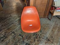 Front view of the orange molded plastic chair seated on a wooden floor with visible wear marks on the seat.