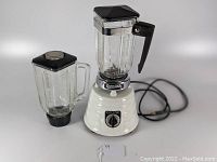 Full view of vintage Osterizer blender with attached glass jar and second jar beside it, black lids on top of jars, white base with ridged design and power cord visible in background.