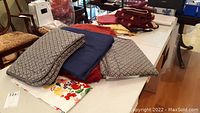 Various linens including folded patterned electric blanket, dark blue fabric (duvet cover), red and cream bath towels, and assorted table linens on flat surface.