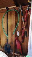 Photo shows two tree loppers hanging on a wooden wall, a green garden hose coiled on a metal holder, and a large blue metal pot.