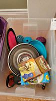 Pet supplies lot contents in clear plastic bin showing food bowls, rubber toys, frisbee, and packaging for Gentle Leader collar