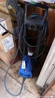 Full view of the Simoniz pressure washer showing blue body, black handle, hose and power cord wrapped around it, and storage location in a garage with boxes.