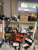 Photo of shelving unit filled with assorted tools, boxes and parts as described by seller, including red and black power tools, boxes and hardware items stacked and loose wiring visible.