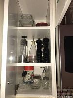 Photo showing clear glass oil and vinegar dispensers, salt and pepper grinders, and small glass jars on a shelf.