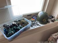 Plastic army toys including soldiers, vehicles, and helmet packed in a plastic container and displayed on a window sill.