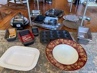 Overview of assorted baking pans, muffin tin, loaf pans, a roasting rack, and ceramic bowls arranged on a rug.