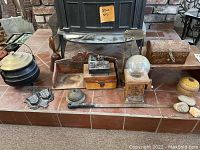 Full view of items arranged on tiled hearth including cast iron kettle, wooden boxes, bell, saber, bubblegum holder, small metal tray with birds, and miniature metal desk scene sculpture.