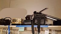 Photo of the shelf showing camera gear including camera bags, tripod, boxed Nikkor lens, and electronics