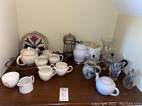 Wide view of the coffee and tea set including white ceramic teapots, sugar and creamer sets, French press, pour-over coffee pot, honey container, and a woven basket