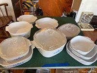 View showing nine white ceramic Corning Ware baking dishes of various sizes and shapes including three with glass lids. Blue floral pattern visible on some dishes.