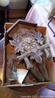 Top view of wooden cuckoo clock in box showing carved leaf details and clock face with Roman numerals, pinecone weights nearby