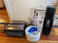 Photo showing the Cuisinart electric griller, immersion blender with box, SodaStream sparkling water maker, and a stack of paper plates
