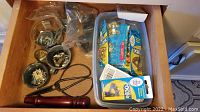Drawer containing various hardware items including metal sheers with black handles, red wooden handle tool, and plastic bins with packaged hardware such as brass hooks and picture hangers.