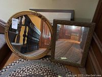 Two gold-tone framed mirrors, oval and rectangular, displayed together leaning on a staircase wall.