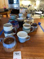 Photo showing a set of eleven pottery style coffee mugs placed on a wooden table in kitchen setting, various colors and shapes visible.