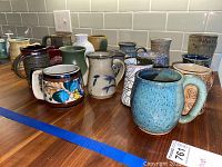 Front view of assortment of pottery coffee mugs showing variety of colors, textures, and shapes on wooden counter with white subway tile background.