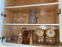 Two clear glass pitchers, multiple glass canisters, two wooden peppermills, and a white ceramic juicer arranged on a wood shelf.