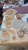 Photo of seashell shaped ceramics and glass plates on white cloth including planter, creamer, sugar dish, and small shell containers