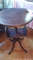 Full view of round wood pedestal table with lower shelf and tripod base, showing general wear and finish.