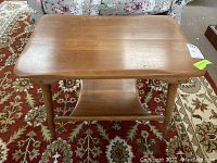 Photo showing solid wood side table from slightly above angle, clear view of tabletop, rounded corners, tapered legs, and bottom shelf, positioned on patterned carpet.