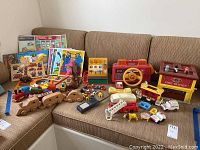 Full view of toys and puzzles arranged on a brown sectional couch including Fisher-Price fire station, wooden puzzles, wooden trains, plastic figures and a red Fisher-Price steering wheel toy.