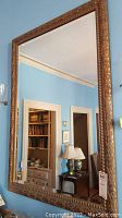 Full view of large rectangular wall-mounted mirror with gold-tone decorative frame against a blue wall showing room reflection.