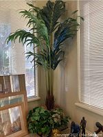 Full view of the artificial palm tree in corner next to window, showing tall fronds and planter base with additional greenery.