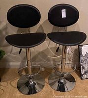 Front view of two black cushioned full backed bar stools with chrome bases and footrests.